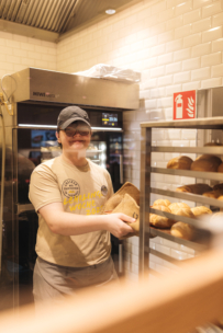 Eine Bäckereimitarbeiterin in Uniform, mit Mütze und Brille lächelt, während sie eine Papiertüte neben einem Regal mit frischen Brotlaiben in einer hellen, modernen Bäckereiküche hält.