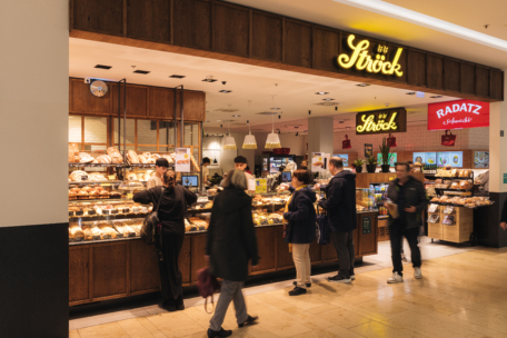 Eine Bäckertheke namens Ströck in einem Einkaufszentrum, in der die Leute bestellen und nach Gebäck suchen. Die Auslage ist mit Backwaren gefüllt, und die Schilder über der Theke sind beleuchtet. Im Hintergrund sind andere Geschäfte und Kunden zu sehen.