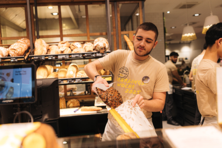 Ein Bäckereiangestellter packt einen Laib Brot hinter einer Theke ein, umgeben von Regalen mit verschiedenen Brotsorten. Er trägt ein beiges Hemd und arbeitet zusammen mit anderen Mitarbeitern in einer hellen, modernen Bäckerei.