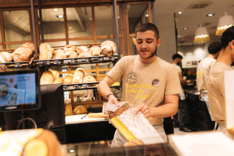 Ein junger Bäckereiangestellter im beigen Hemd legt lächelnd einen Laib Brot in eine Papiertüte. Hinter ihm befinden sich Regale mit verschiedenen Brotsorten, und im Hintergrund ist ein weiterer Mitarbeiter zu sehen.