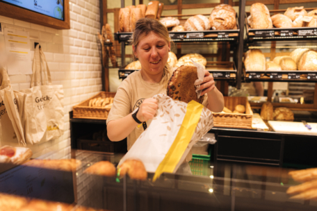 Eine Frau in einer Bäckerei legt einen großen Laib Brot in eine Papiertüte hinter einer mit Backwaren gefüllten Glastheke, mit Regalen mit verschiedenen Brotsorten im Hintergrund.