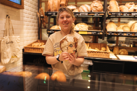 Eine Bäckereimitarbeiterin steht hinter dem Tresen, lächelt und hält einen Laib Brot in der Hand. Im Hintergrund sind Regale mit verschiedenen Brotsorten zu sehen. Die Umgebung ist warm und einladend.