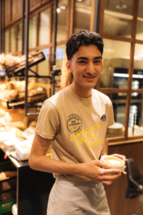 Ein junger Mann mit beigem T-Shirt und Schürze lächelt, während er eine Tasse Kaffee in einer Bäckerei in der Hand hält. Im Hintergrund sind Regale mit Brot und Gebäck zu sehen.
