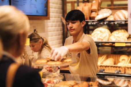 Eine Bäckereimitarbeiterin mit Handschuhen serviert einem Kunden an einer Bäckertheke Gebäck, während im Hintergrund Regale mit verschiedenen Brotsorten und Backwaren zu sehen sind. Ein weiterer Mitarbeiter arbeitet hinter der Theke.