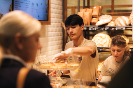 Ein Bäckereiarbeiter mit Handschuhen serviert einem Kunden ein Gebäck, während ein anderer Mitarbeiter in der Nähe arbeitet. Im Hintergrund sind Regale mit verschiedenen Brotsorten zu sehen.