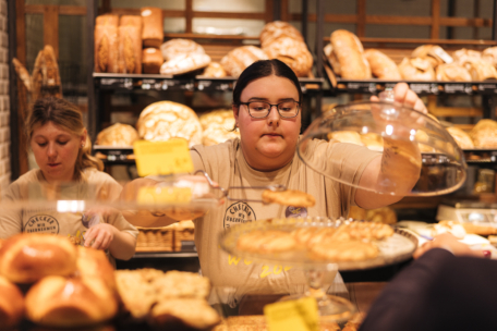 Zwei Personen arbeiten hinter einer Bäckertheke; die eine legt Gebäck auf ein Tablett unter einer Glaskuppel, während die andere daneben steht. Im Hintergrund sind Regale mit verschiedenen Brotsorten zu sehen.