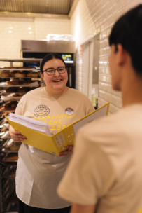 Eine lächelnde Frau mit Brille und Schürze hält einen großen gelben Ordner in der Hand, während sie sich mit einer anderen Person in einer Bäckereiküche unterhält; im Hintergrund sind Brotstangen zu sehen.