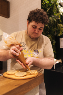 Eine Person mit lockigem Haar benutzt einen Spritzbeutel, um Kekse auf einem Holzbrett zu dekorieren. Sie trägt ein beiges Hemd und ein Namensschild und steht in einer Küche mit weißen Kachelwänden und Grünpflanzen im Hintergrund.