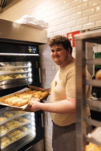 Eine Person steht in einer Bäckereiküche, lächelt und hält ein Tablett mit frisch gebackenem Gebäck vor einem Ofen, in dem weiterer Teig auf Tabletts aufgeht. Im Hintergrund sind weiß gekachelte Wände und Metallgestelle zu sehen.