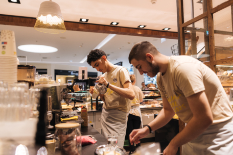 Zwei Baristas in beigen Hemden arbeiten hinter einer geschäftigen Café-Theke und bereiten Getränke und Speisen zu. Im Hintergrund stehen Regale mit Gebäck und Brot, und das Café ist von warmem Licht und Menschen erfüllt.