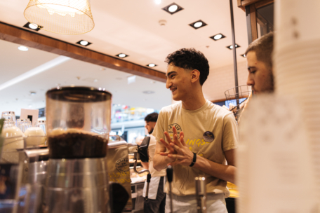 Ein Barista lächelt und unterhält sich mit einer Mitarbeiterin hinter einer Kaffeetheke in einem hell erleuchteten Café, mit Kaffeegeräten und Tassen im Vordergrund.