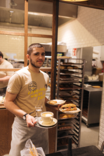 Ein junger Mann in einem beigen T-Shirt und einer Schürze steht in einer Bäckereiküche und hält Teller mit einer Tasse Kaffee, einem Keks und Gebäck in der Hand. Im Hintergrund sind Regale mit Backwaren und ein weiterer Arbeiter zu sehen.
