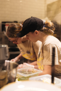 Zwei Personen in hellbraunen Hemden, schwarzen Mützen und Handschuhen bereiten gemeinsam in einer Küche Essen zu und konzentrieren sich dabei auf ihre Arbeit. Um sie herum sind verschiedene Küchenbehälter und -utensilien zu sehen.