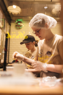 Zwei Angestellte mit Haarnetzen, Handschuhen und beigen T-Shirts bereiten hinter einem Tresen in einer Restaurantküche mit weißen Kachelwänden und heller Beleuchtung Speisen zu.
