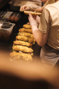 Eine Person mit Handschuhen stellt Sandwiches auf einer schwarzen Theke zusammen und legt das obere Brötchen auf die Reihen der vorbereiteten und mit Grünzeug belegten Sandwiches. Im Hintergrund sind andere Lebensmittel zu sehen.