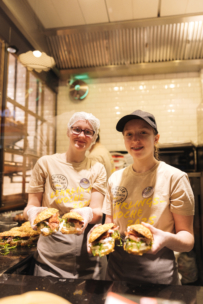 Zwei lächelnde Gastronomieangestellte in beigen Hemden und Hüten stehen hinter einer Theke und halten große Sandwiches mit verschiedenen Zutaten in einem gut beleuchteten Sandwich-Laden.