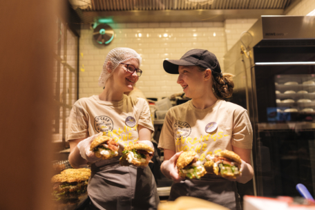 Zwei lächelnde Angestellte in Uniformen und mit Hüten halten große Burger in einer Restaurantküche. Sie stehen nebeneinander, einander zugewandt, mit weiß gekachelten Wänden und einem Kühlschrank im Hintergrund.