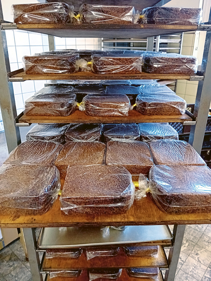 Auf mehreren Regalböden eines Bäckereiregals stehen ordentlich gestapelte, quadratische Schokoladenkuchen, die in Plastikfolie eingewickelt sind, bereit zur Lagerung oder Auslieferung. Der Hintergrund zeigt eine geflieste Wand und weitere Backbleche.