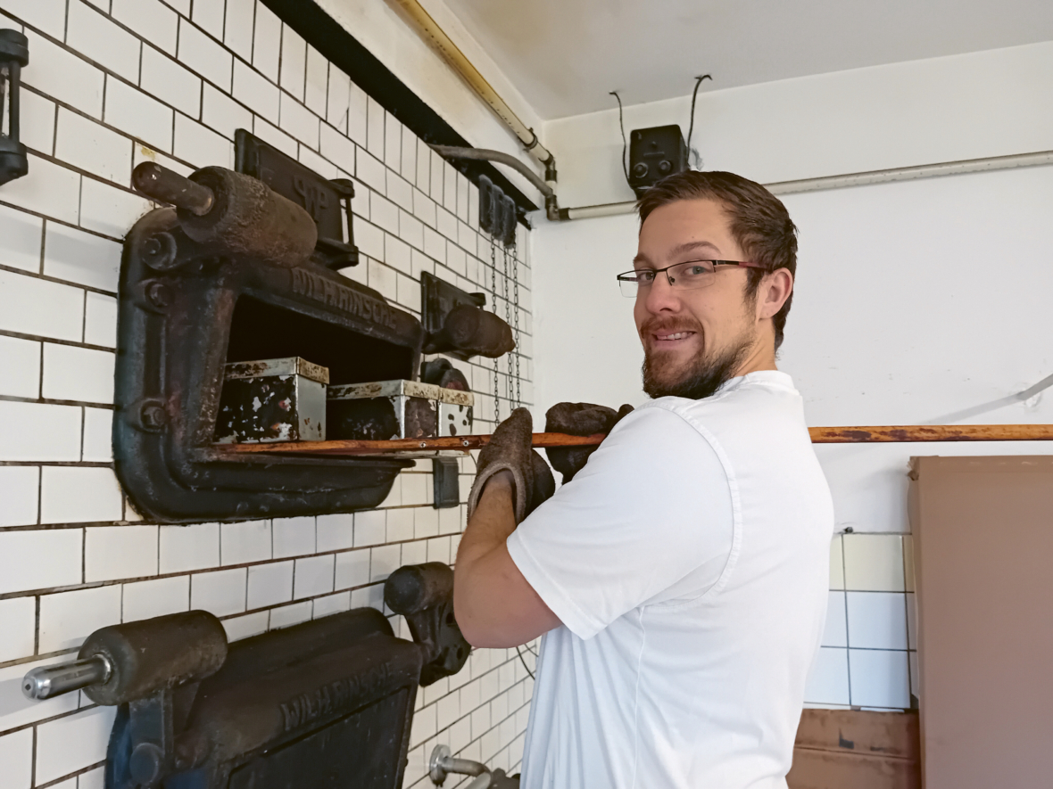 Ein Mann mit Brille und Handschuhen lächelt in die Kamera, während er ein langes hölzernes Werkzeug benutzt, um etwas aus einem alten Ofen in einer gekachelten Küche zu entfernen.