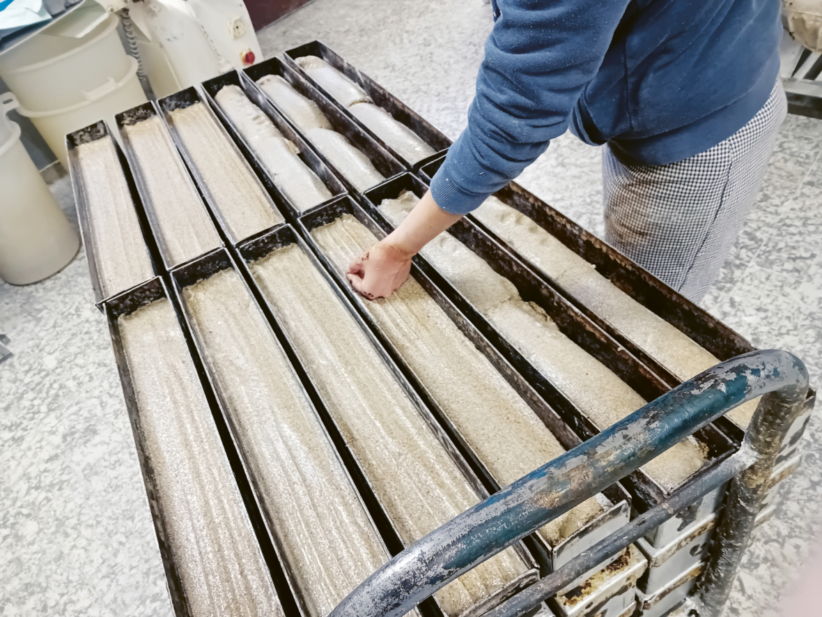 Eine Person streut Mehl über mehrere rechteckige Brotteige, die in Metallschalen auf einem Wagen angeordnet sind, wahrscheinlich in einer Bäckerei.