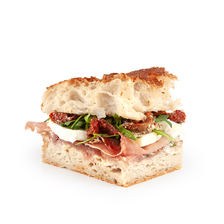 Eine Nahaufnahme eines rustikalen Sandwiches mit Prosciutto, Mozzarella, Rucola und sonnengetrockneten Tomaten auf knusprigem Brot, isoliert auf weißem Hintergrund.