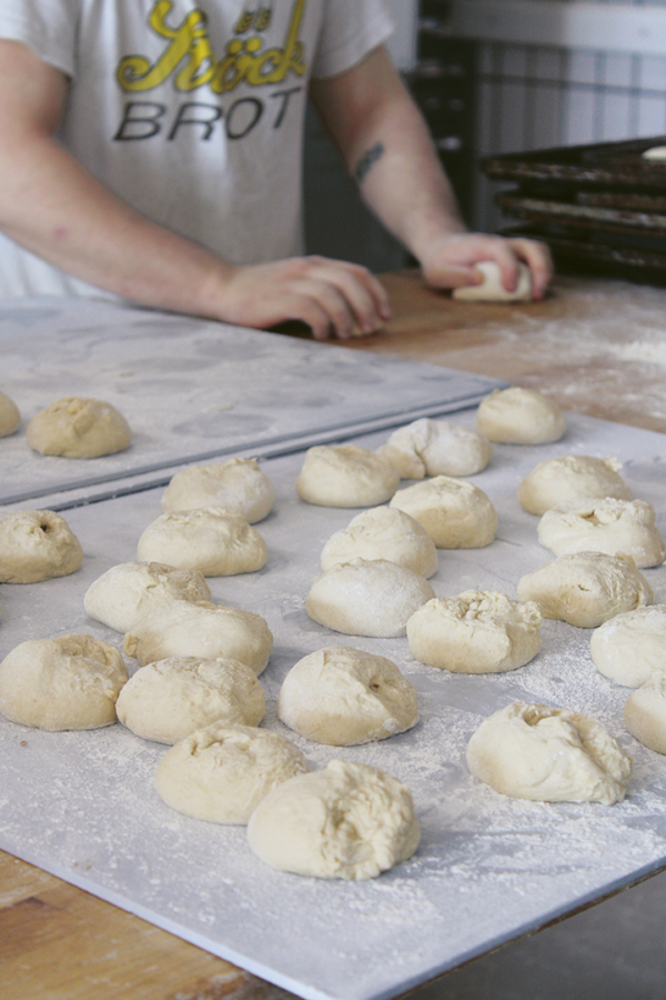 Eine Person formt Teig auf einer bemehlten Holzfläche, wobei viele kleine Teigkugeln auf zwei Blechen im Vordergrund ruhen, bereit zum Backen. Auf dem Hemd der Person steht "Stück BROT.".