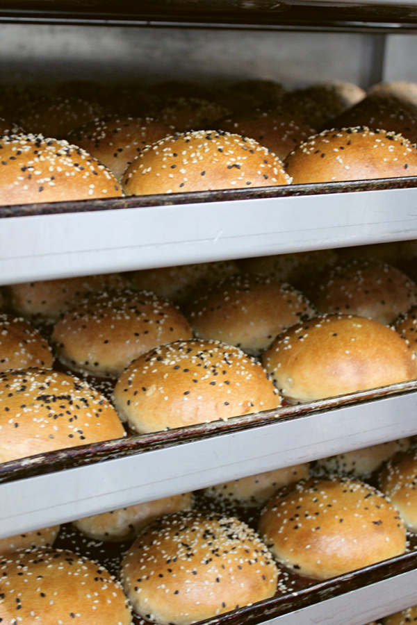 Reihen von goldbraunen, mit Sesam und Mohn belegten Hamburgerbrötchen kühlen auf Metalltabletts in einem Bäckereigestell ab.