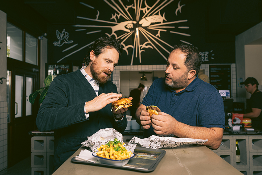 Zwei Männer stehen drinnen, untersuchen und halten Burger, mit Pommes und Essen auf einem Tablett vor ihnen. Im Hintergrund sind künstlerische Wanddekorationen und eine warme, einladende Beleuchtung in einer zwanglosen Restaurantumgebung zu sehen.