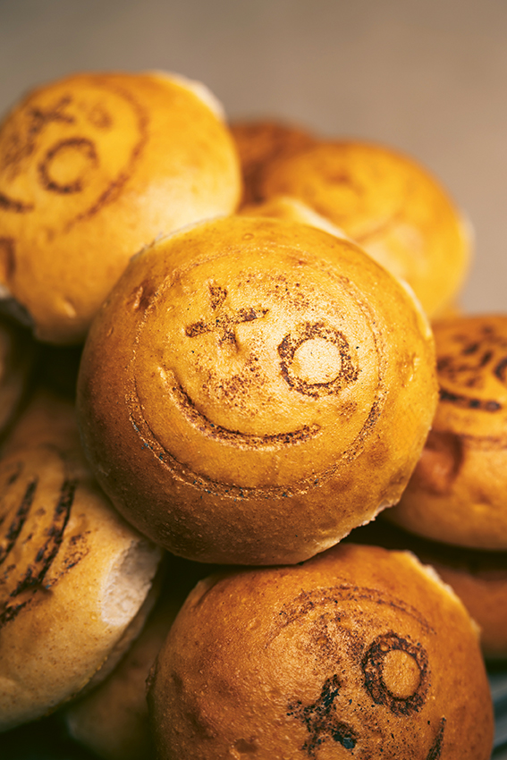 Eine Nahaufnahme von runden Brötchen, die übereinander gestapelt sind. Auf jedem Brötchen ist ein lächelndes Gesicht mit den Symbolen X und O eingeprägt. Die Brötchen sind goldbraun und scheinen frisch gebacken zu sein.