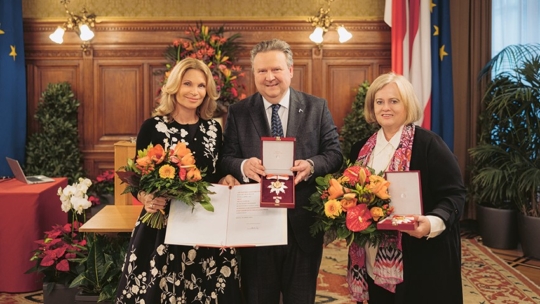 Drei Personen stehen in einem Haus und halten Medaillen, Urkunden und Blumensträuße in der Hand. Sie sind förmlich gekleidet, lächeln und posieren vor einer Holzvertäfelung, Fahnen und Blumenarrangements, die eine Preisverleihung andeuten.
