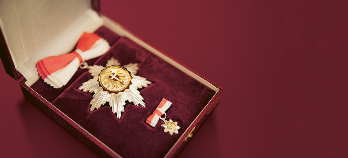Ein dekoratives Medaillenset mit einem goldenen und weißen sternförmigen Abzeichen, einer rot-weißen Bandschleife und einer kleineren, passenden Anstecknadel, die alle in einer mit Samt ausgekleideten Präsentationsbox auf weinrotem Hintergrund präsentiert werden.