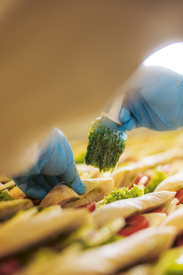Eine Person mit blauen Handschuhen streicht eine grüne Kräutermischung auf die mit Salat, Tomaten und anderen Zutaten belegten Sandwiches. Das Bild ist eine Nahaufnahme, die sich auf den Zubereitungsprozess konzentriert.