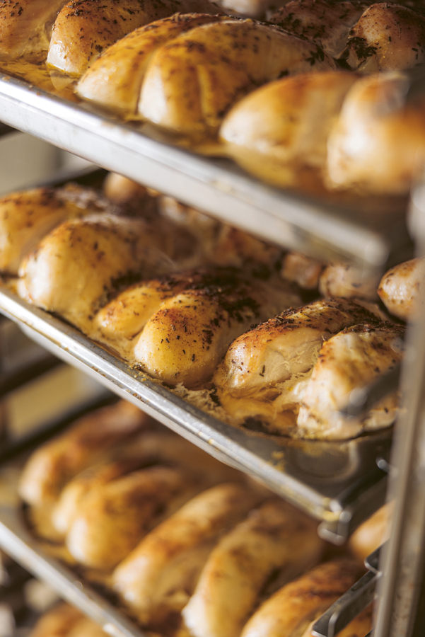 Reihen von goldgelben, gewürzten Brathähnchen, die auf Metalltabletts in einer Großküche oder einem Backofengestell ruhen, mit aufsteigendem Dampf und einem warmen, appetitlichen Aussehen.