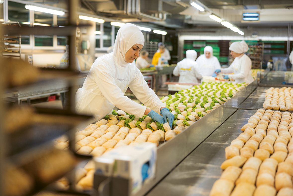Eine Frau, die einen weißen Hidschab, Handschuhe und eine Uniform trägt, bereitet an einer großen Bäckereifertigungsstraße Gebäck zu, während im Hintergrund andere Arbeiter in ähnlicher Kleidung arbeiten. Reihen von ungebackenem Gebäck sind fein säuberlich auf Tabletts angeordnet.