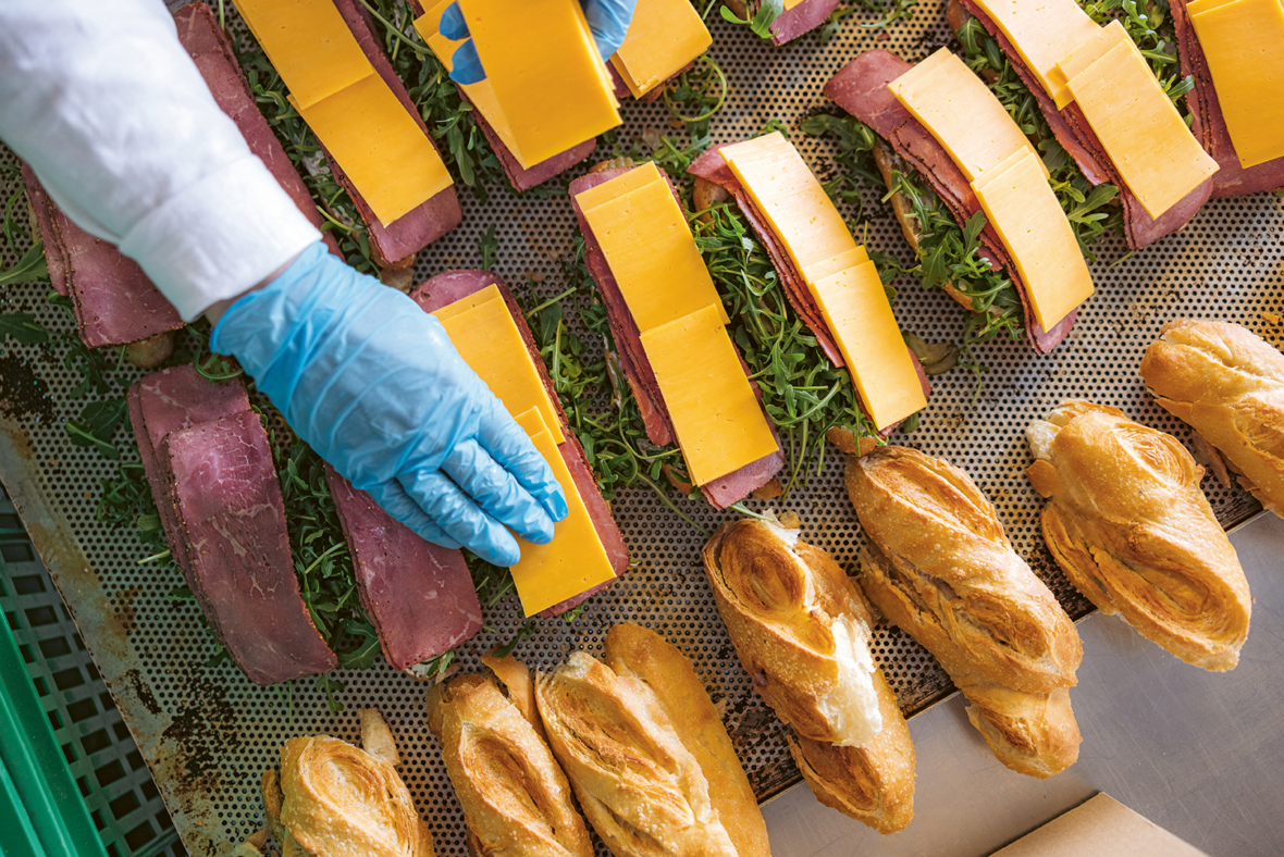 Eine Person mit blauen Handschuhen legt Cheddar-Käsescheiben auf mit Fleisch und Gemüse gefüllte Sandwiches neben einer Reihe frischer Baguettes auf eine perforierte Metallfläche.
