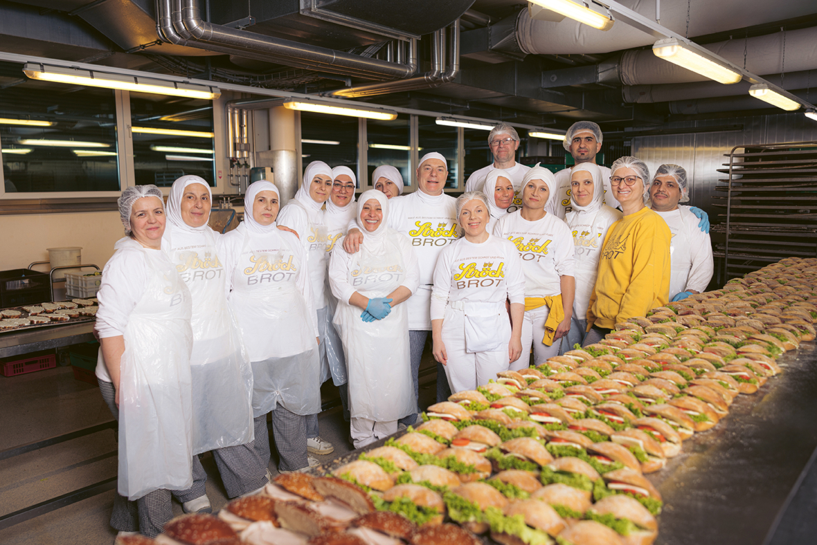 Eine Gruppe von Bäckereiangestellten in weißen Uniformen und Haarnetzen posiert gemeinsam in einer Küche und steht hinter einer langen Reihe ordentlich belegter Sandwiches mit Salat, Käse und Tomaten.