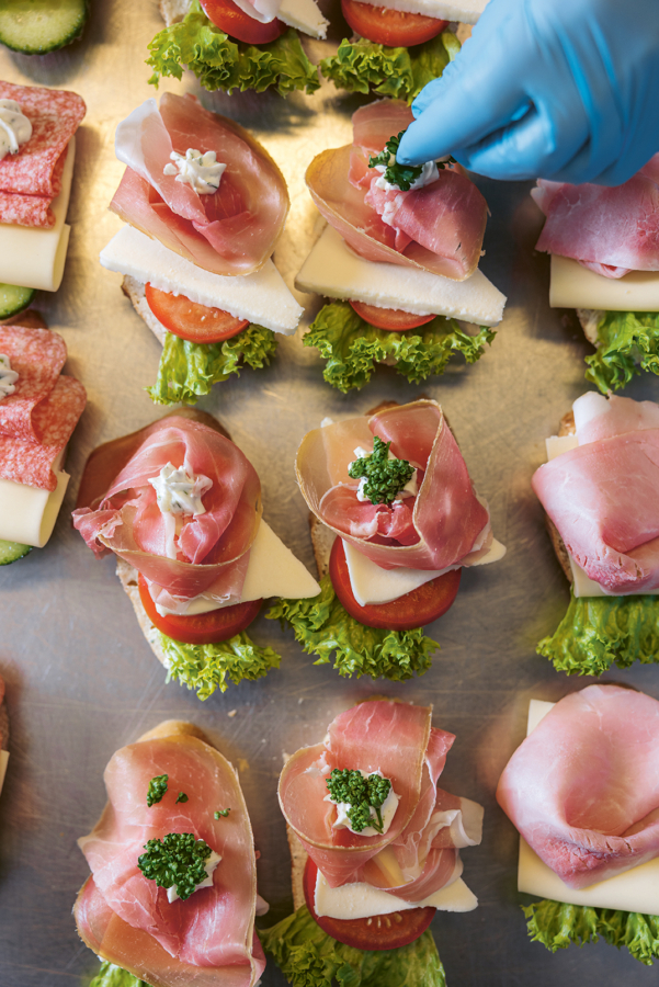 Auf einem Tablett sind belegte Brötchen mit Salat, Tomaten, Käse und dünnen Scheiben gepökelten Fleisches angerichtet. Eine behandschuhte Hand garniert eines der Sandwiches mit Petersilie.