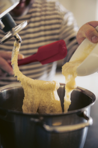 Eine Person kratzt mit einem roten Spatel klebrigen Teig aus einem Standmixer-Aufsatz, während sie Teig aus einer weißen Schüssel in eine große Metallschüssel gießt. Die Person trägt ein gestreiftes Hemd.