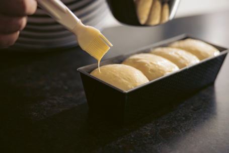 Mit einem Backpinsel wird der aufgegangene Teig in einer rechteckigen Laibform mit Ei oder Butter bestrichen, um ihn für das Backen auf einer dunklen Arbeitsfläche vorzubereiten.