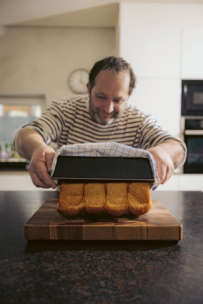 Ein Mann in einem gestreiften Hemd lächelt, während er in einer modernen Küche einen Laib Brot aus einer Backform auf ein hölzernes Schneidebrett stürzt.