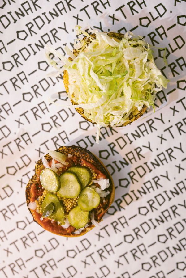 Ein offener Hamburger mit zerkleinertem Salat auf dem oberen Brötchen und Essiggurken, Zwiebeln und Soße auf dem unteren Brötchen, platziert auf einem Papier mit der Aufschrift "XTRA ORDINARY" in schwarzer Schrift.