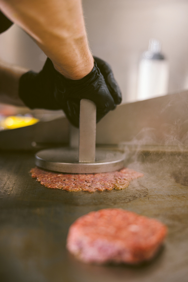 Eine Person, die einen schwarzen Handschuh trägt, drückt eine runde Burgerpresse aus Metall auf rohes Rinderhackfleisch auf einer heißen Grillplatte, wodurch das Fleisch beim Garen flachgedrückt wird. Dampf steigt von der Grillfläche auf.