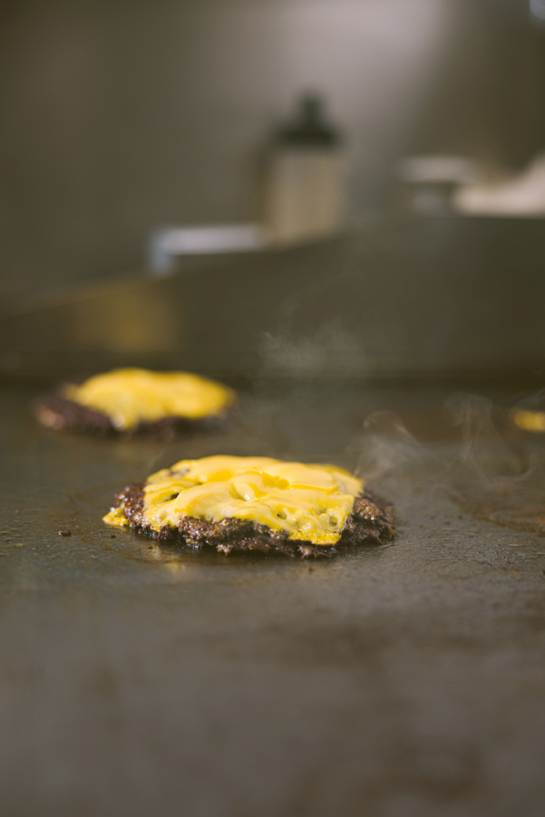 Eine Nahaufnahme von zwei Rinderpasteten mit geschmolzenem Käse, die auf einer heißen, dampfenden Grillplatte zubereitet werden, mit einem verschwommenen Küchenhintergrund.