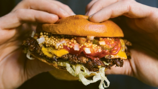 Nahaufnahme von Händen, die einen Hamburger mit getoastetem Brötchen, geraspeltem Salat, geschmolzenem Käse, Essiggurken, gewürfelten Zwiebeln, Ketchup, Senfkörnern und einer saftigen Rindfleischpastete halten.