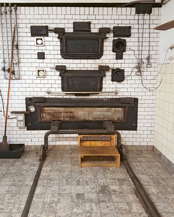 Große Industrieöfen mit schwarzen Metalltüren und Drehknöpfen sind in eine weiß gekachelte Wand eingebaut. In der Nähe hängen Metallwerkzeuge, und vor den Öfen steht ein kleiner hölzerner Trittschemel auf einem gefliesten Boden mit Metallschienen.