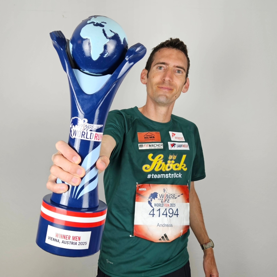 Ein Mann in grünem Hemd und Startnummer hält eine große blaue Trophäe mit einer Weltkugel auf der Spitze. Auf der Trophäe steht WINNER MEN VIENNA, AUSTRIA 2023. Er lächelt und steht vor einem schlichten Hintergrund.