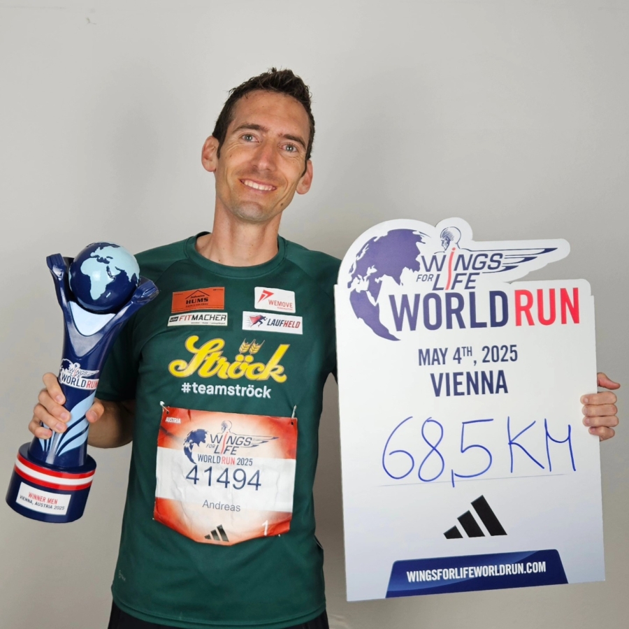 Ein lächelnder Mann in einem grünen Hemd hält eine Trophäe in der einen und ein Schild in der anderen Hand, auf dem "Wings for Life World Run, 4. Mai 2025, Wien, 68,5 km" steht. Auf seiner Startnummer steht der Name Andreas.