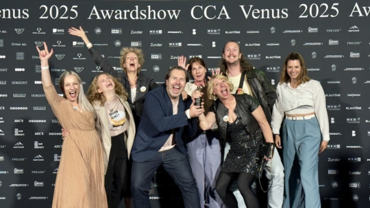 Eine fröhliche Gruppe von acht Personen posiert gemeinsam auf einem roten Teppich bei der CCA Venus 2025 Awardshow, hält eine Trophäe in der Hand und lächelt aufgeregt vor einem Hintergrund, der mit Sponsorenlogos bedeckt ist.