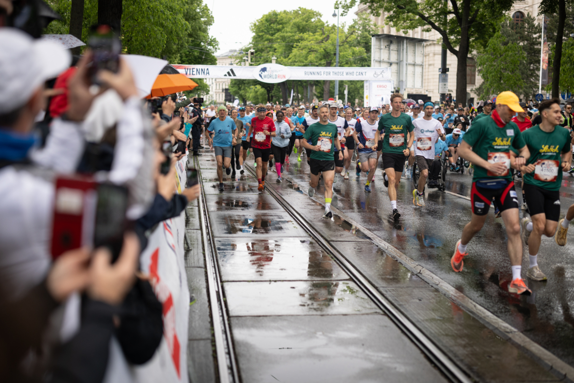 Eine große Gruppe von Läufern in Sportkleidung nimmt an einem regnerischen Tag an einem Stadtlauf teil. Die Zuschauer halten Regenschirme und jubeln am Rande der nassen Straßenbahnschienen und Straßen.