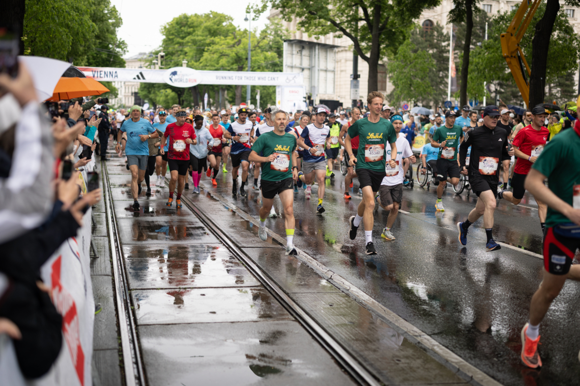 Läuferinnen und Läufer nehmen an einem verregneten Tag an einem Stadtmarathon teil. Die Straßen sind nass und die Zuschauer jubeln ihnen von der Seite zu. Viele Läuferinnen und Läufer tragen grüne Hemden und Startnummern, während sie auf einer Straßenbahnstrecke unter grünen Bäumen laufen.